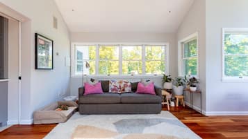 A spacious living room with a grey sofa, a patterned rug, and large windows offering a view of trees.