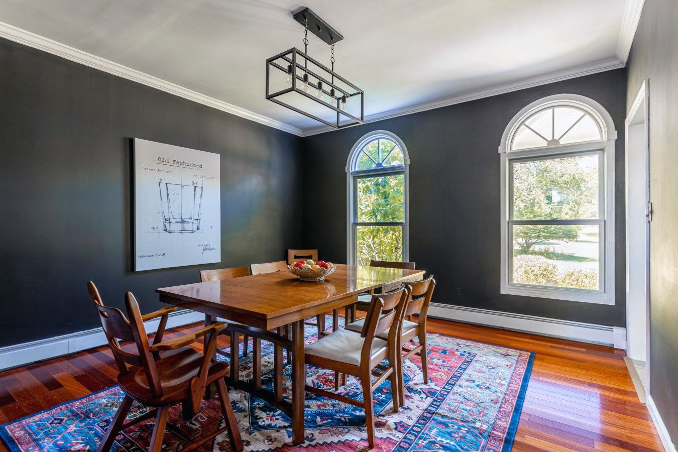 A dining room with a wooden table, chairs, a chandelier, a large window, and a framed architectural drawing on the wall.
