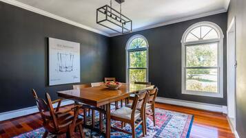 A dining room with a wooden table, chairs, a chandelier, a large window, and a framed architectural drawing on the wall.