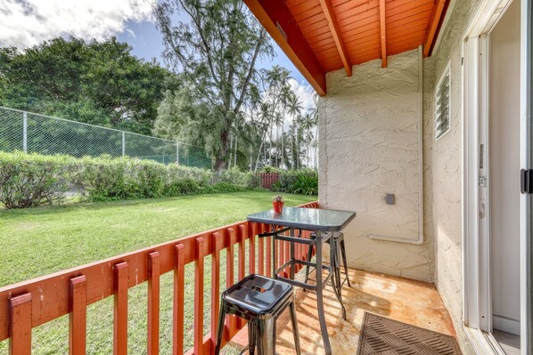 Un espacio al aire libre techado con una mesa y sillas, baranda roja, y vista a un jardín con árboles.