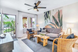 Una sala de estar moderna con un sofá, dos sillones de mimbre, una mesa de centro, un ventilador de techo y un ventanal con cortinas.