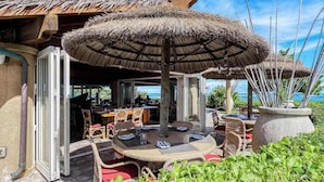 An outdoor dining area with round tables, thatched umbrellas, wicker chairs, and a view of the ocean. - NEW Fall Deal Water-Slide Resort Days at Oceanfront Waipouli (Kapaa)