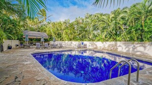 Piscina com piso de pedra, rodeada por palmeiras e uma área de estar coberta.