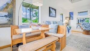 A room with a wooden bench, a table, a bowl of flowers, and a view of palm trees through the window. - NEW Corner Princeville Loft Studio with Hanalei Mountain Views (Princeville)