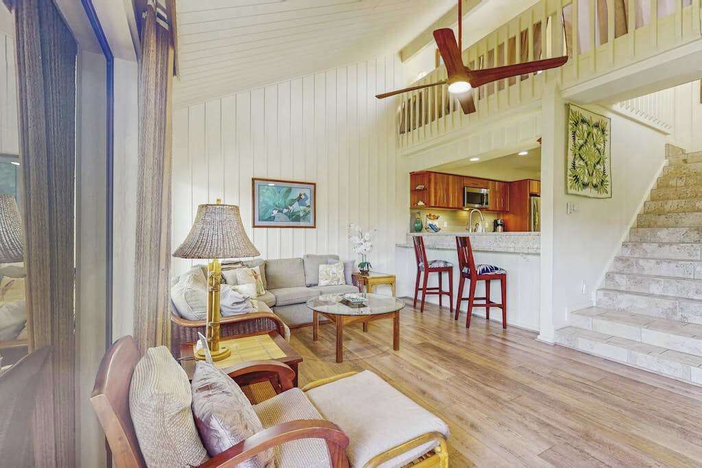 A spacious living area with a sofa, chairs, a coffee table, and a dining table. A kitchen is visible in the background, and a staircase leads to an upper level.