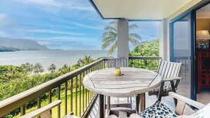 A balcony with a round wooden table, two chairs, a vase, and a view of the ocean and mountains. - *NEW* Fall Special Two King Suites with Wide Hanalei Bay Views (Princeville)