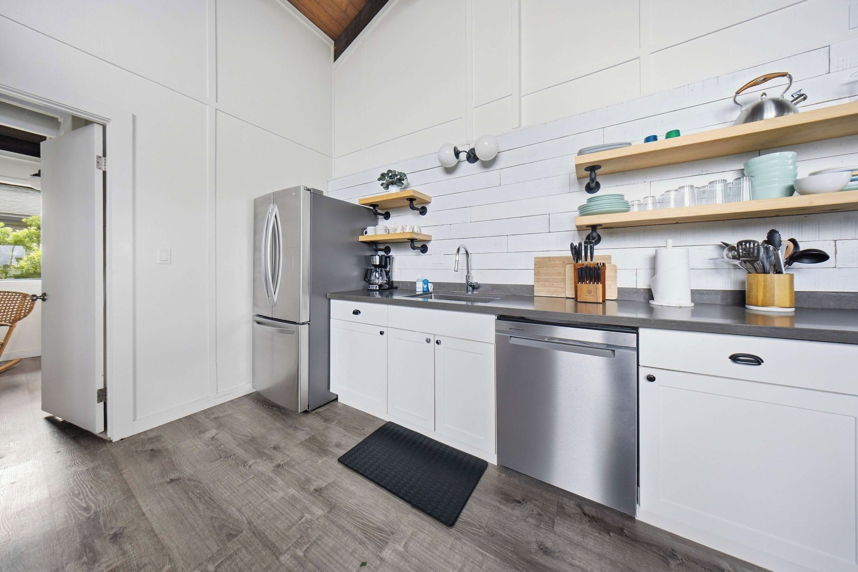 A modern kitchen with white cabinetry, a stainless steel refrigerator, and a dishwasher. There are wooden shelves above the countertop holding dishes and utensils.