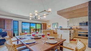 A dining area with a wooden table set for four, a kitchen with modern appliances, and large windows offering a view. - *New* Oceanfront Wailua Escape Near Lae Nani Beach (Kapaa)