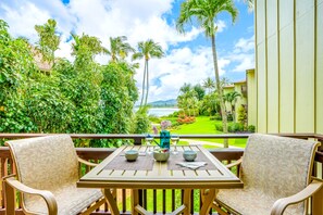 A balcony with two chairs and a table, overlooking a lush garden and a view of the ocean. - *NEW* Fall Special Beachfront Kapaa Condo for Sunrise Walks and Pool Afternoons (Kapaa)