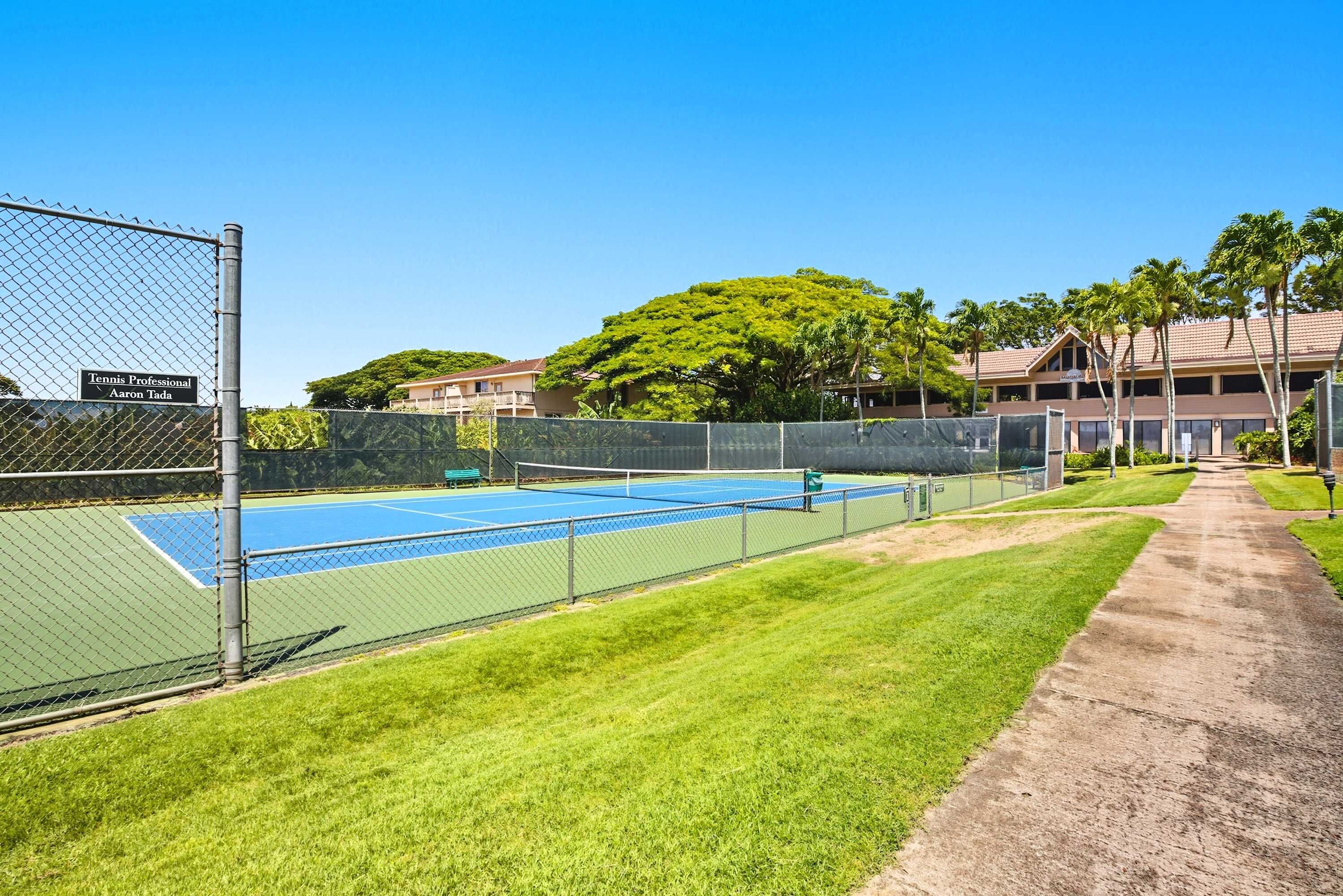 A tennis court with a chain-link fence, a sign for 'Tennis Professional Aaron Tada', and a pathway leading to a building with a red roof.