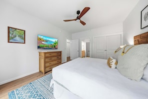 Ein Schlafzimmer mit einem Bett, einem Fernseher, einem Deckenventilator, einem Schminktisch und einer Tür zu einem anderen Raum.