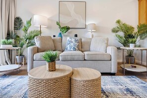 A living room with a grey sofa, two round woven side tables, a blue patterned throw pillow, and several potted plants. - Poipu Mermaid Family Condo - Pool, Hot Tub, Tennis, AC, Kitchen, Laundry, Beach (Koloa)