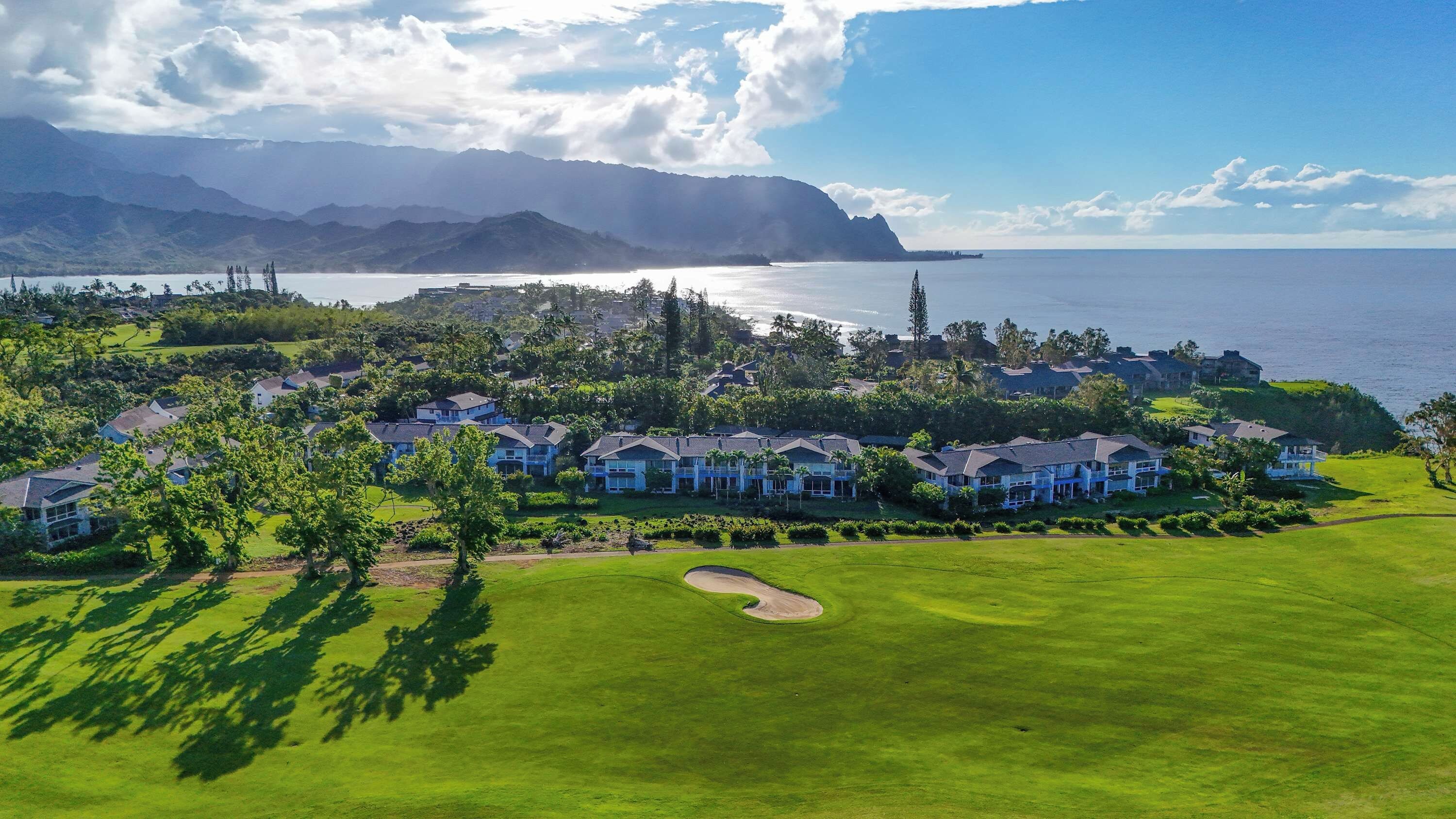 Un campo de golf con un club de campo y edificios residenciales, rodeado de vegetación exuberante y con vistas a un cuerpo de agua y montañas al fondo.