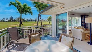 Ein Balkon mit Blick auf einen Golfplatz, Korbmöbel und eine Glastür zu einem Innenbereich mit einer Couch und einer Lampe.