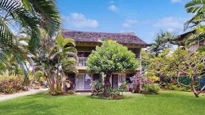 Una casa de dos pisos con balcón, rodeada de césped cuidado y vegetación tropical.