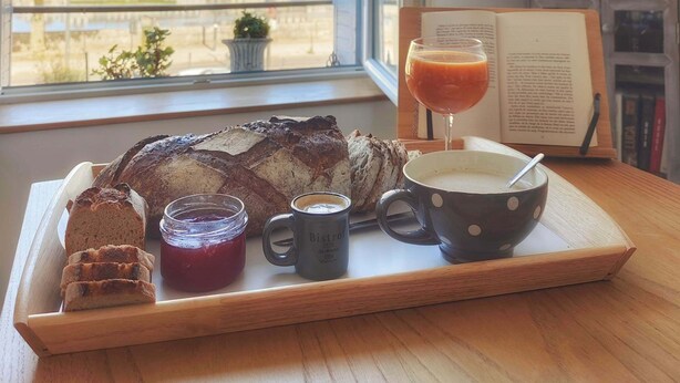 Een ontbijttafel met brood, jam, koffie en een drankje, bij een raam met uitzicht op een stadsgezicht.