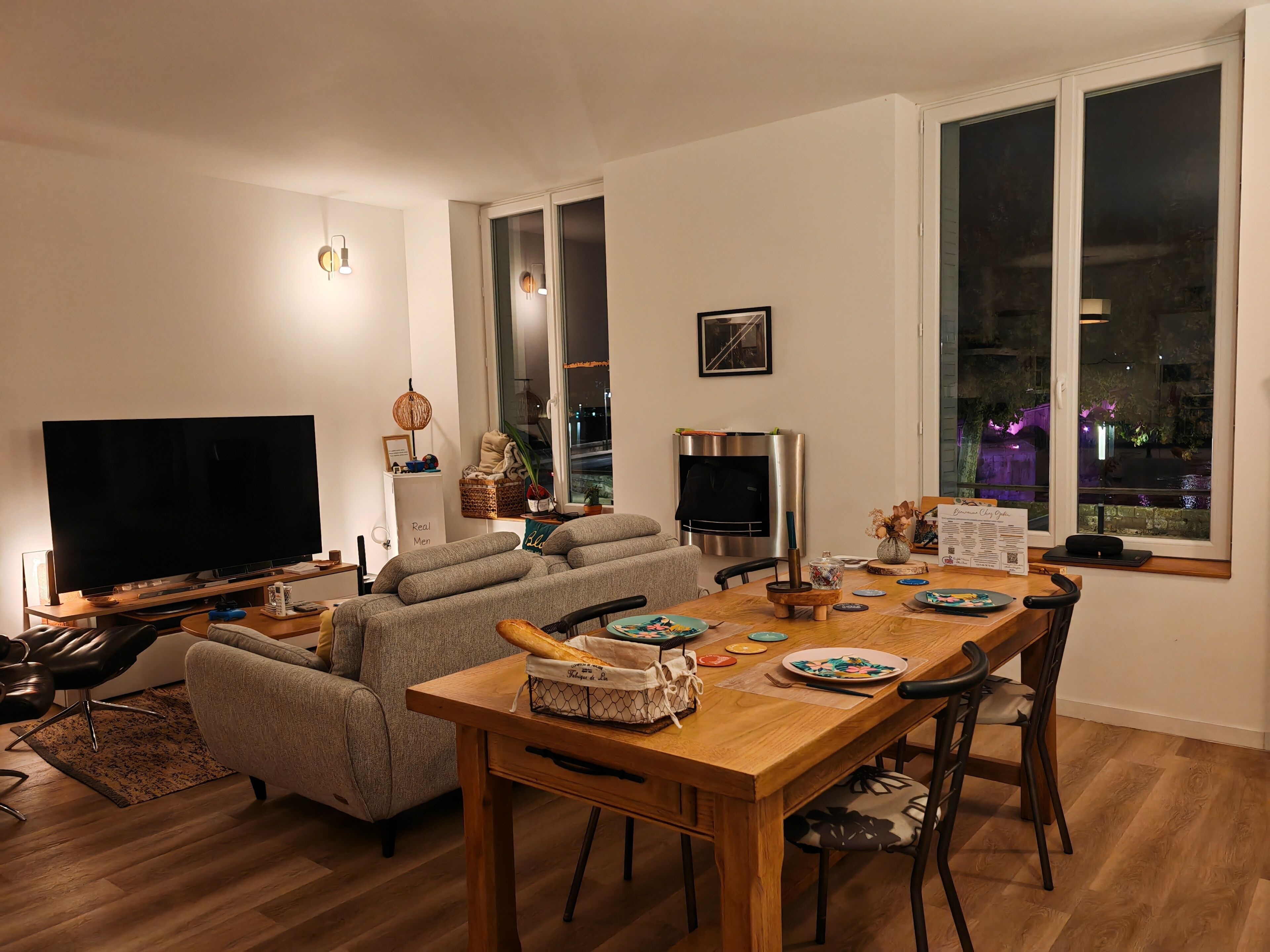 A modern living room with a flat-screen TV, a sofa, a dining table with chairs, and a large window with a view of the outside.