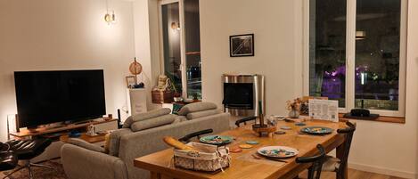 A modern living room with a flat-screen TV, a sofa, a dining table with chairs, and a large window with a view of the outside.