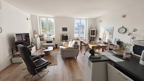 Een moderne woonkamer met een bank, salontafel en televisie. Er is een eethoek met een tafel en stoelen. De kamer heeft grote ramen met uitzicht op gebouwen en bomen.