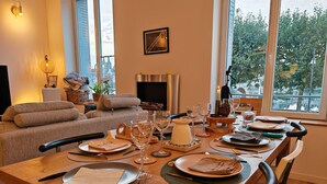 Een eethoek met een lange tafel gedekt voor vier personen, een bank, een open haard en uitzicht op een stadsbeeld door grote ramen.