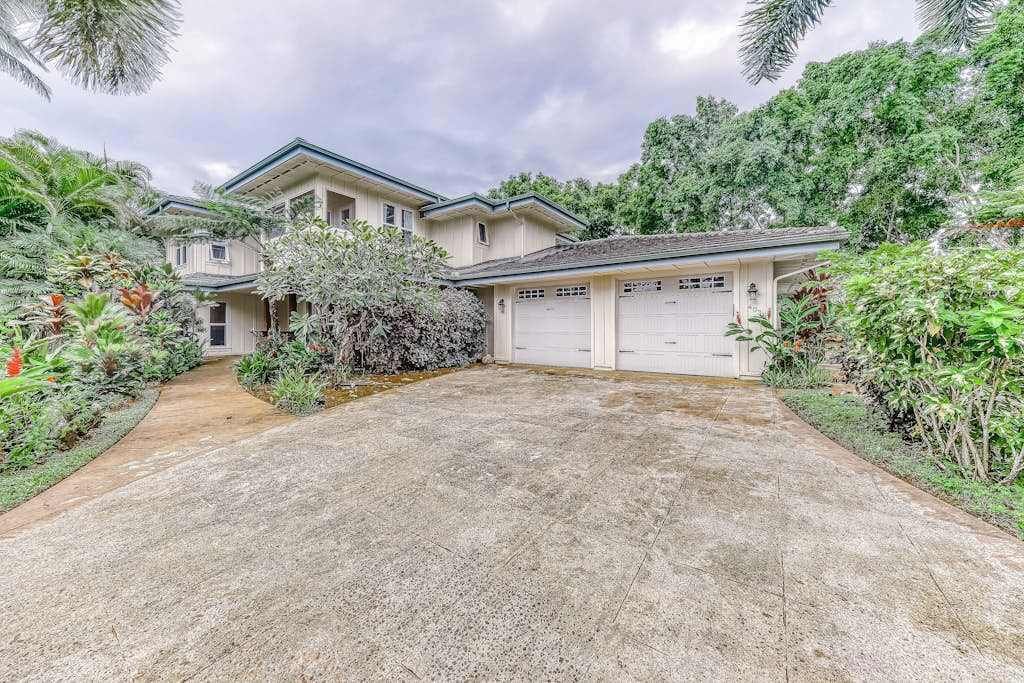 A two-story house with a garage, surrounded by lush greenery and a concrete driveway.