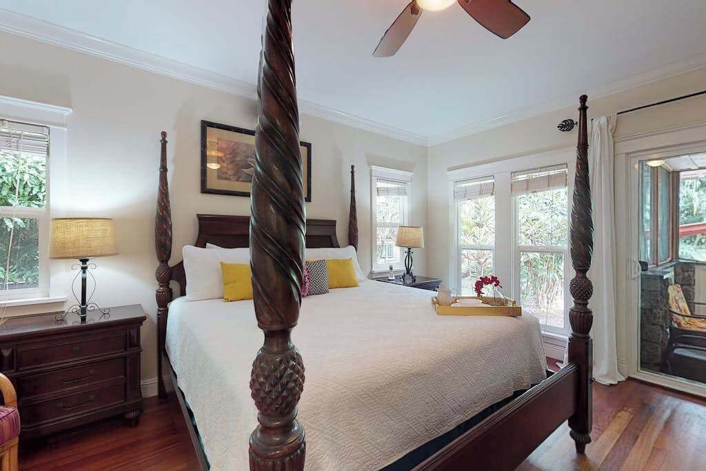 A four-poster bed with a wooden headboard, a nightstand with a lamp, and a window with a view of greenery.