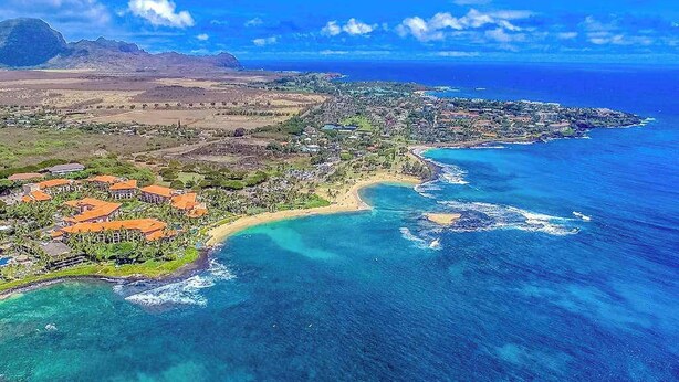 An aerial view of a coastal area with a beach, clear blue waters, and a mountainous backdrop. - *NEW* Air-Conditioned Poipu Retreat, 100 Yards from Brennecke's (Koloa)