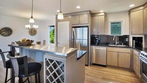 A modern kitchen with stainless steel appliances, a central island with bar stools, pendant lighting, and a view of greenery outside. - *NEW* Princeville Ocean Views for Slow Kauai Mornings (Princeville)