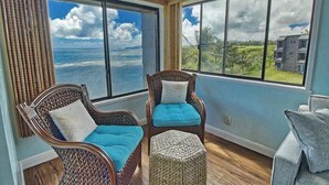 Una habitación con ventanas amplias, sillas de mimbre con cojines, un reposapiés tejido y una lámpara.