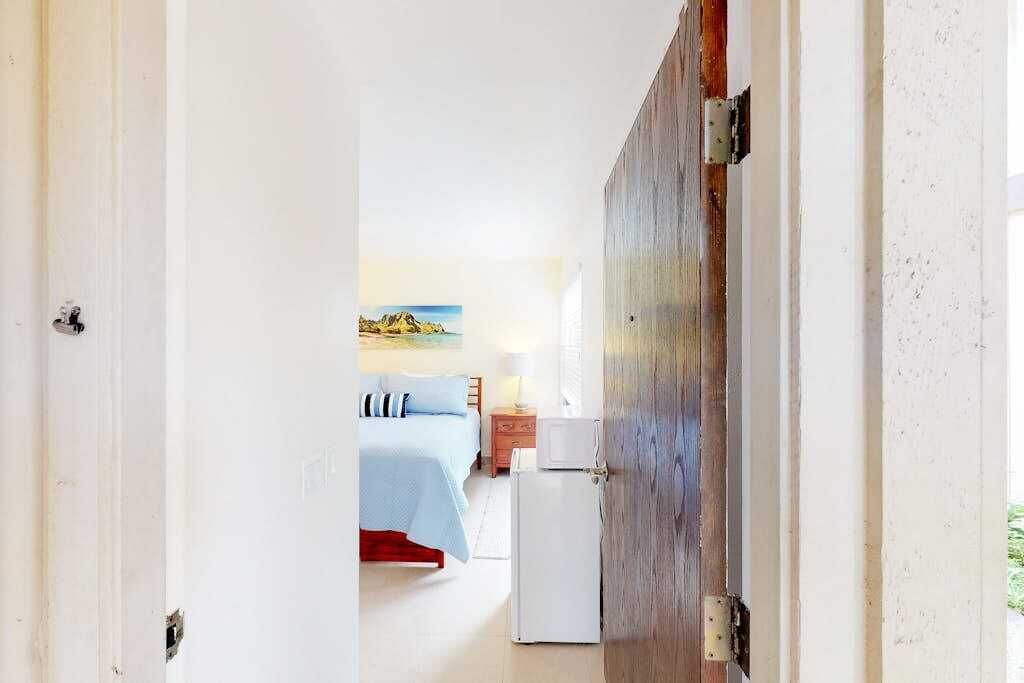 Uma porta que dá para um quarto com uma cama, mesa de cabeceira e um refrigerador.
