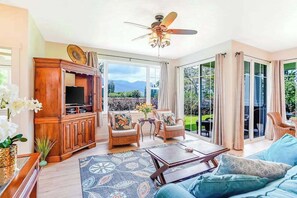 A spacious living room with a television, wicker furniture, a ceiling fan, and large sliding glass doors leading to a garden.