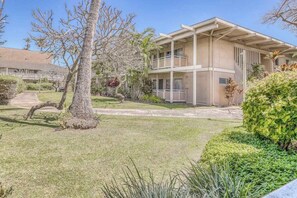 A two-story building with a flat roof, a covered patio, and a well-maintained lawn with trees and shrubs. - Ground-Floor Plantation Hale Bungalow-AC, Pools, Spa, WIFI, Shops, Dining, Beach (Kapaa)