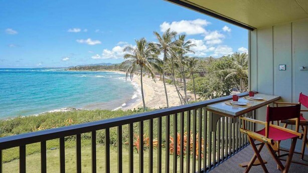 A balcony with a view of a beach and ocean, a table with chairs, and a railing. - *NEW* Beachfront Kapa'a Stay with Pool and Coastal Views (Kapaa)