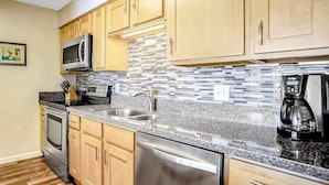 Una cocina con gabinetes de madera, lavavajillas de acero inoxidable, microondas, cafetera y salpicadero de azulejos.