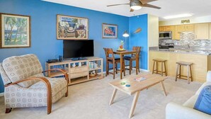 Una sala de estar con televisor, muebles de madera y una zona de cocina con mostrador y alacenas.