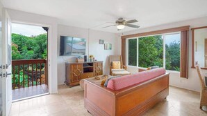 Une chambre avec un lit, une télévision, un ventilateur de plafond, une fenêtre donnant sur un espace verdoyant et un balcon.