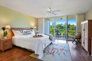 Quarto de hotel com cama, mesa de cabeceira, abajur, janela com vista para vegetação e ventilador de teto.