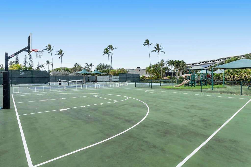 Ein Basketballplatz im Freien mit Korb, umgeben von Palmen und einem Spielplatz.