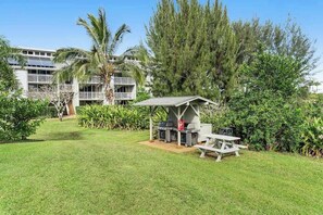 A grassy outdoor area with a barbecue setup and a picnic table, surrounded by lush greenery and a building with balconies. - NEW North Shore Sunrises and Bali Hai Sunsets in Princeville (Princeville)