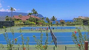 En tennisbane med net, omgivet af grønt og med et bjerg i baggrunden.