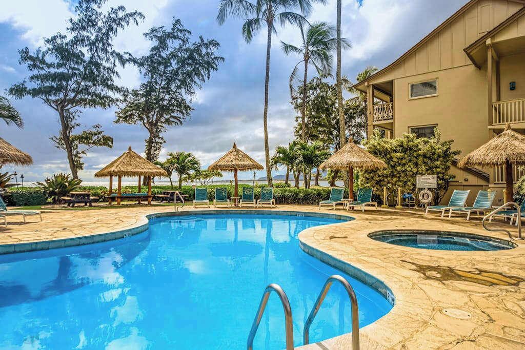 A pool area with lounge chairs, thatched umbrellas, and a building with balconies.
