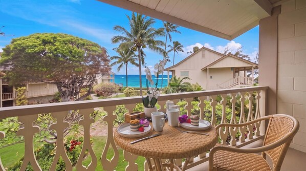 Un balcon avec vue sur l’océan, une table à manger avec des chaises et des plantes tropicales.