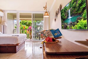 Une chambre d’hôtel avec un lit, une table à manger avec un ordinateur portable, un téléviseur diffusant une image de cascade et un balcon agrémenté de plantes.