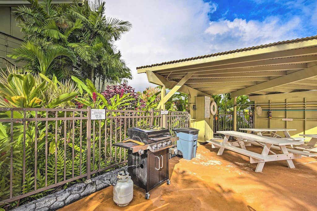 Un espacio al aire libre cubierto con una parrilla, mesas de picnic y un jardín cercado con plantas tropicales.