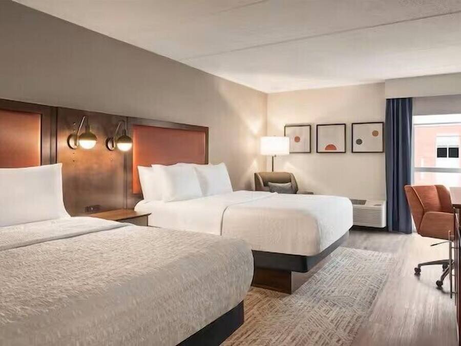 A hotel room with two beds, a desk, a chair, a TV, and artwork on the wall.
