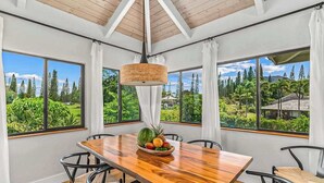 Uma área de refeições com mesa de madeira, cadeiras, uma fruteira e janelas amplas com vista para o verde.