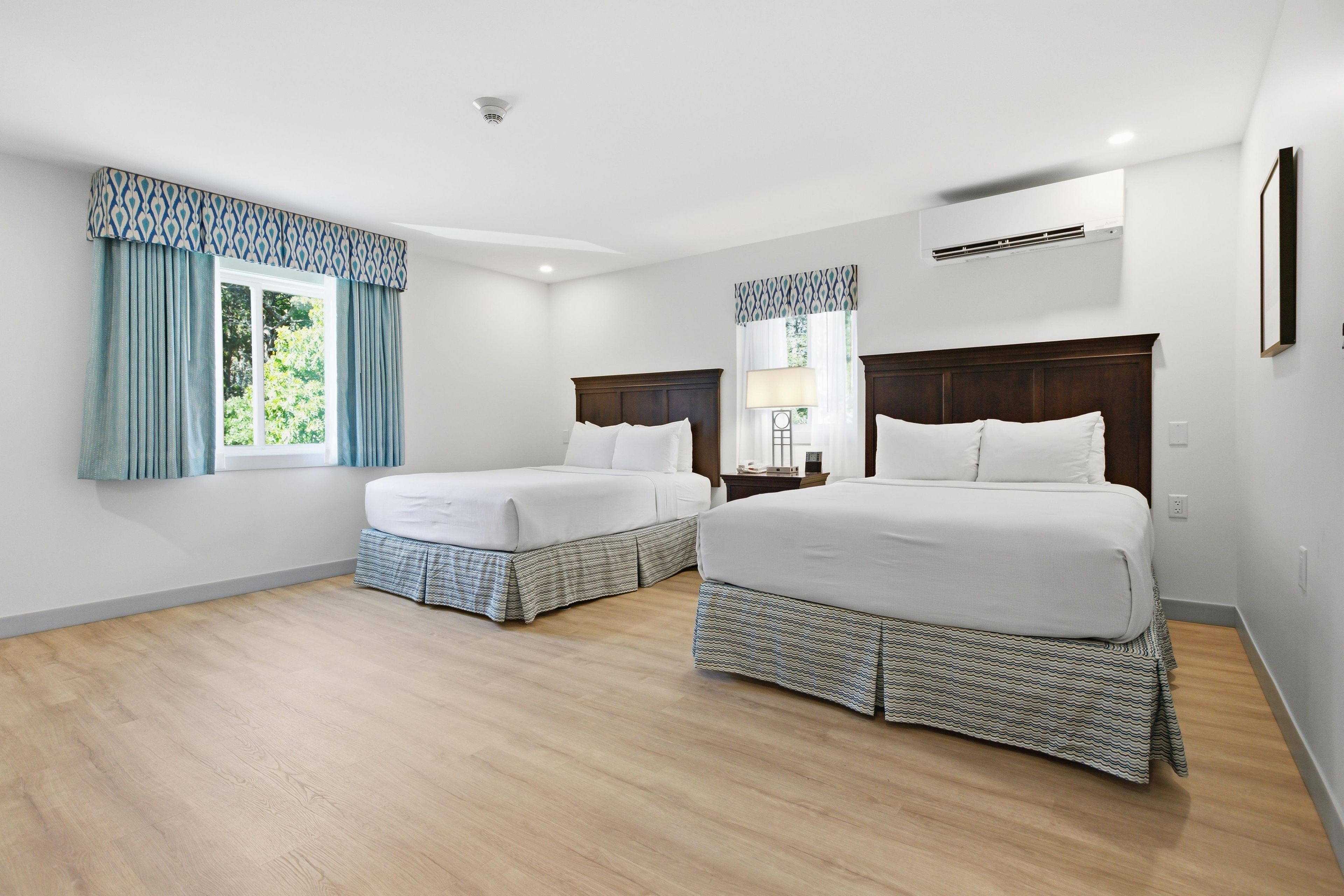 Une chambre d’hôtel avec deux lits, une fenêtre avec des rideaux, une table de chevet avec une lampe et un appareil de climatisation fixé au mur.