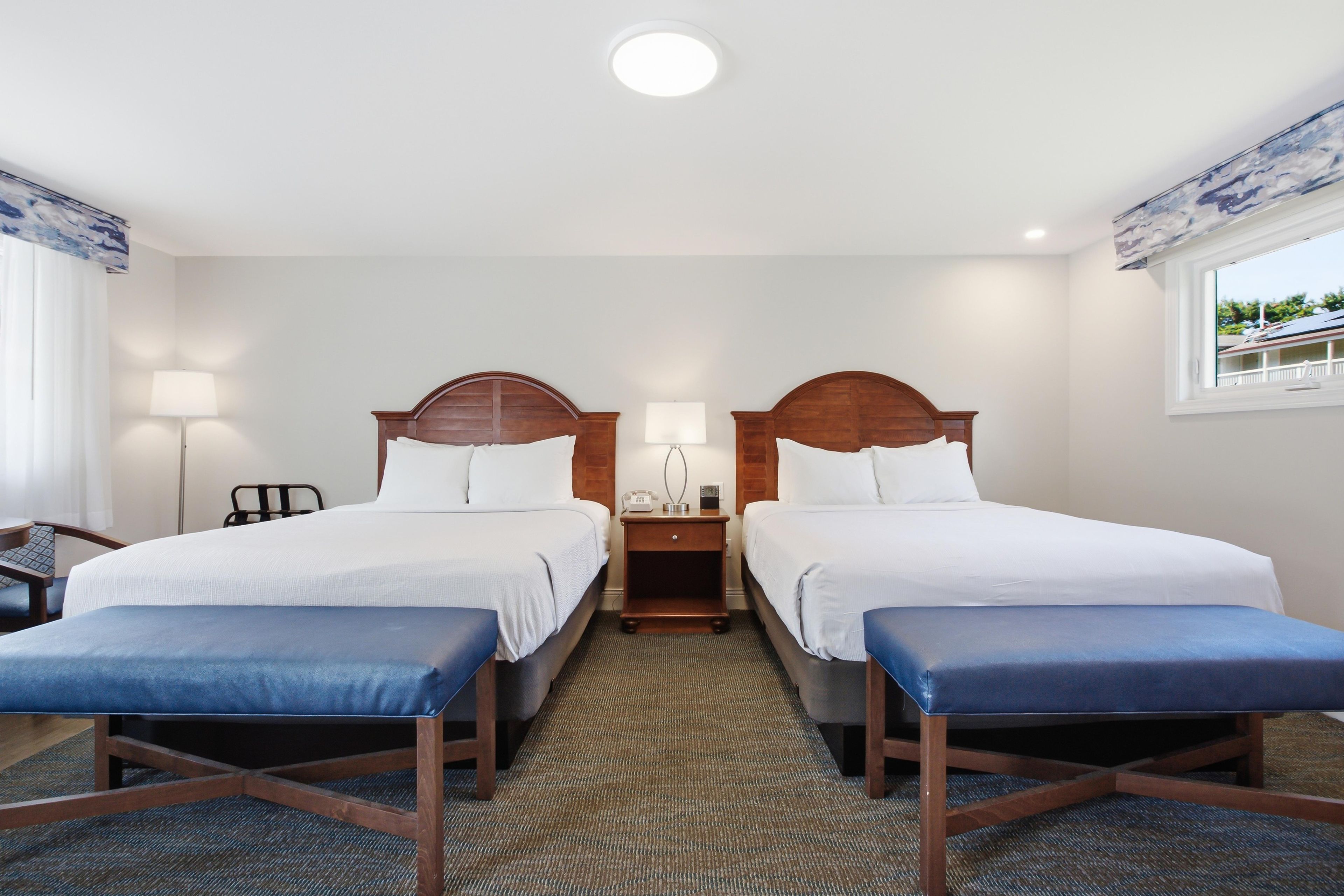 Quarto de hotel com duas camas, uma escrivaninha, uma cadeira, um abajur e uma janela.