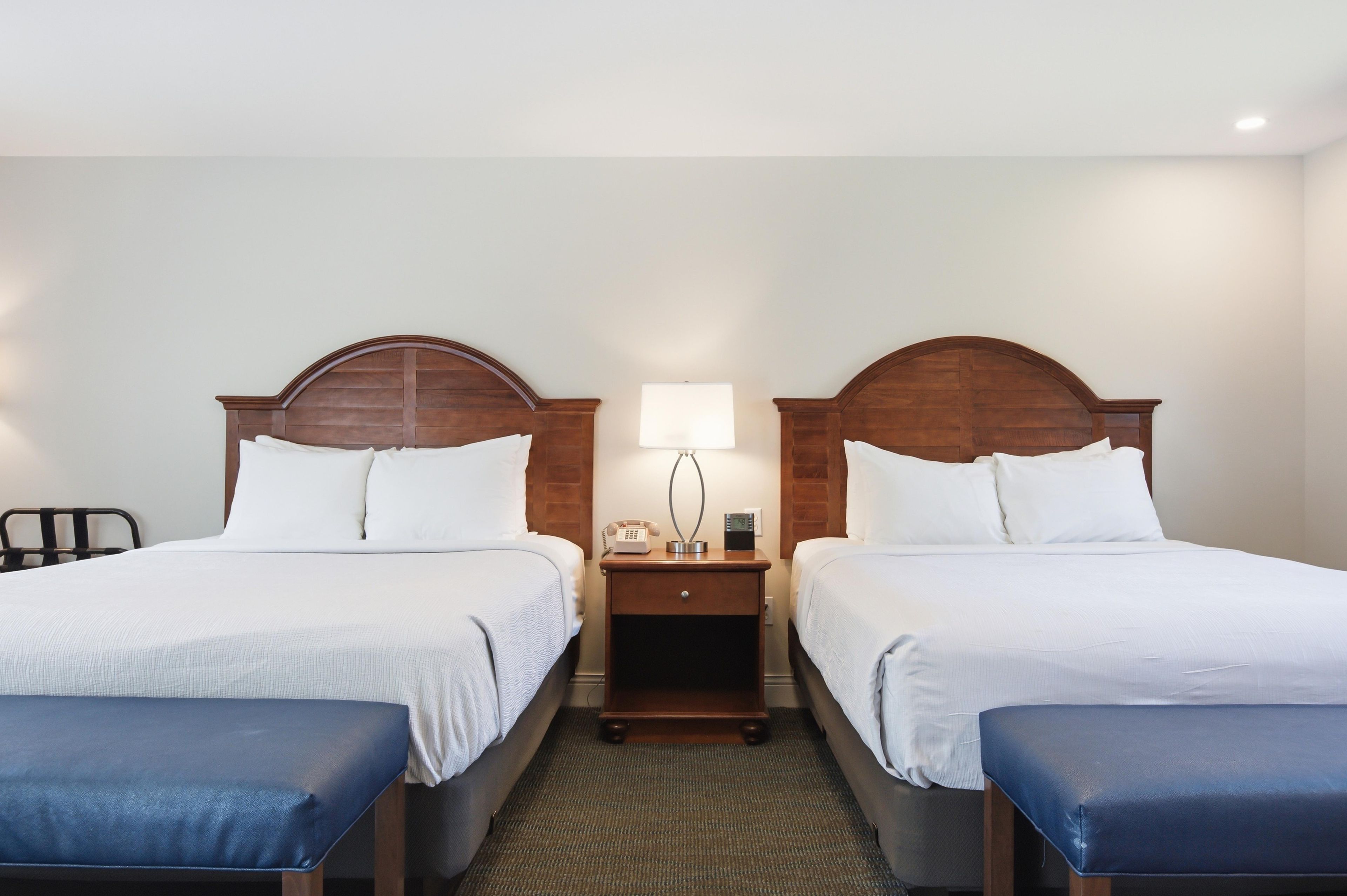 A hotel room with two beds, a nightstand with a lamp, and a chair.