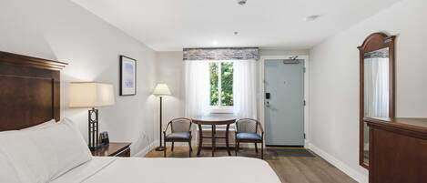 A hotel room with a bed, two chairs, a table, a desk, a mirror, and a door leading to another room.
